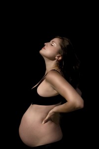 Pregnant female with a burnished gold and copper tint to her skin wearing a black bra and pants and set against a black background.